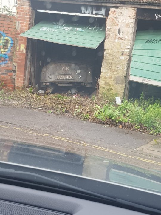 'Barn find' but what is it? - Page 1 - General Gassing - PistonHeads UK - This image depicts a car parked in an outdoor area that appears to be under a roof or shed. The vehicle is old and rusted, suggesting it has been exposed to the elements for some time. It's situated on a roadway with a green patch of ground around it, possibly grass or moss growth. In the background, there are structures partially obscured by rainwater, indicating recent precipitation. To the right, there is a building with graffiti visible on its wall, and to the left, a portion of another car can be seen. The scene has an urban, gritty feel, accentuated by the overcast sky and the wet conditions on the ground.