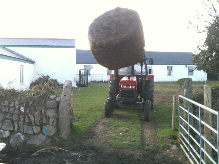 An old truck is parked on the side of the road - Pistonheads - The image shows a narrow dirt road with a rustic brown stone wall lining one side. A red tractor with a large square bale of hay attached to the back is parked along this road, indicating agricultural activity. There's another tractor in the background, but it's much smaller and less significant than the primary one. Buildings can be seen in the background, suggesting a rural or semi-rural residential or farming area. The lighting and shadows suggest it's midday with a clear sky.