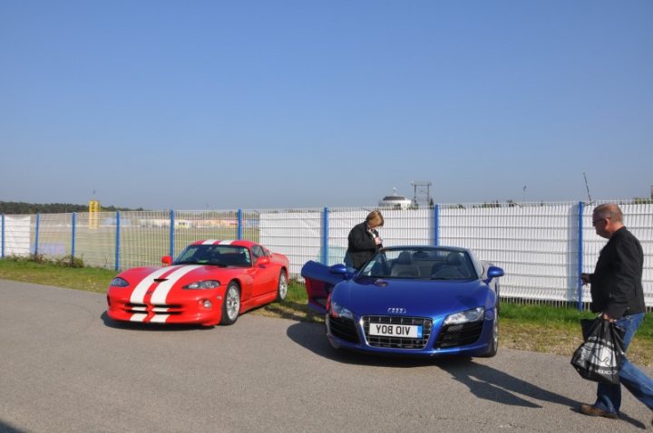 Jim Clark Hockenheimring Pistonheads - The image shows a scene of a car sale lot. Two cars are parked parallel to each other, one is a striking red with white stripes, suggesting it might be a sports car or a convertible. The other car is a shade of blue and sports the Audi logo. There are two individuals in the image; one is right in front of the camera, appearing to look at the viewer, and the other is farther away, near the red car, seemingly engaged in a conversation or transaction. The environment suggests an outdoor setting with a paved area, a fence in the background, and no individuals currently interacting directly with the cars.