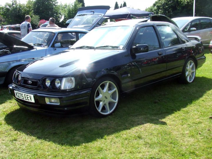 South Wales run and BBQ 21st Aug - Page 1 - South Wales - PistonHeads - The image displays a black sedan, likely a Ford, parked on a grassy surface. It has a glossy finish on the body and shiny rims on the wheels. The car appears to be in sunlight, as indicated by the shadows and reflections visible on the car's surface. In the background, there are other vehicles, including a hard-top convertible, suggesting this could be a car show or a gathering of vehicle enthusiasts. The focus is on the front end of the black sedan, which is turned slightly towards the camera, allowing a clear view of its grille, headlights, and one side of the car.