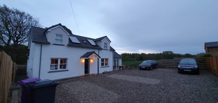 Black / Grey Fascia With White Windows And Render - Page 1 - Homes, Gardens and DIY - PistonHeads - The image captures a scene at dusk. It shows a house with white walls and a gray roof, situated in a rural setting. A car is parked on the gravel driveway leading to the house. The sky above is dark, suggesting either late evening or an approaching storm. There are trees visible in the background, indicating some open space around the property. The image is taken from a perspective that shows both the front of the house and part of its side view.