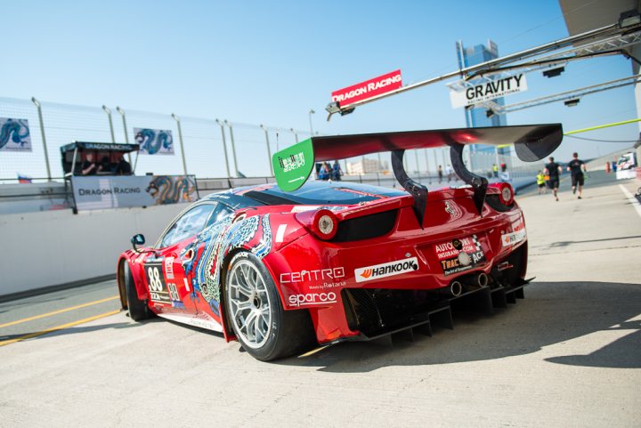 Dubai 24hr Race - Autodrome this weekend - Page 1 - Middle East - PistonHeads - The image captures a red race car on a race track, painted with various designs and logos, highlighting its involvement in a racing event. A rear wing characteristic of racing cars is installed on the car, suggesting it's preparing for a time-to-beat or gravity session. A pit stop service trailer is visible in the background, hinting at logistical support for the racing team. There are spectators in the stands and other cars and trucks in the vicinity, adding to the atmosphere of a racing event.