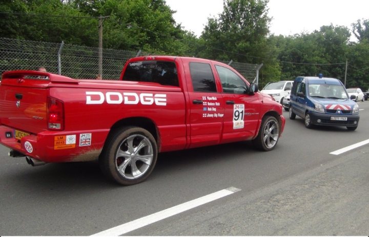 RAM at tunnel for Lemans trip - Page 1 - Vipers - PistonHeads - The image depicts a street scene with various vehicles. In the foreground, there is a red Dodge truck with a white tag that reads "112 89". This truck is adorned with several stickers on its windows and rear section. Behind the Dodge truck, there is a blue and white striped van. Other vehicles are visible as well, indicating that this is a busy section of the road. The road itself appears to be lined with trees and fencing on the sides. The sky is overcast, suggesting that the weather might be cloudy or gloomy.