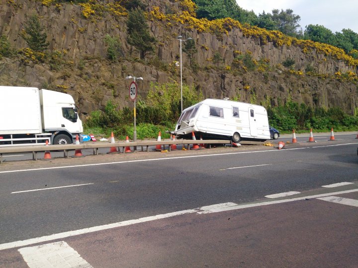 User error - check your hitch! - Page 1 - Tents, Caravans & Motorhomes - PistonHeads - The image shows a scene of an accident on the side of a road. A white camper van is flipped on its side, resting on the guardrail of a road barrier. It has been involved in a collision with another vehicle, which appears to be a white truck. Several orange traffic cones are strategically placed around the accident site, likely to indicate to oncoming traffic to slow down or change lanes. The vehicles are located on a road that is adjacent to a rocky wall or hill, covered in lush greenery and shades of yellow, suggesting the season could be spring or summer. The sky above is cloudy and the overall atmosphere is relatively calm, despite the unusual situation.