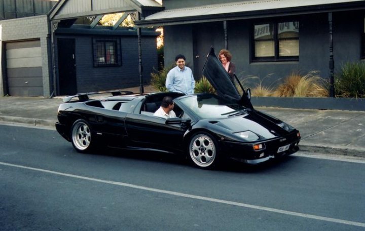 Show us your Diablo  - Page 5 - Diablo/Murcielago/Aventador - PistonHeads - The image captures a scene on a street where a man is seen getting into a black sports car with a white roof. Another person is standing beside the open car door, and there is a third individual slightly behind them, wearing a suit jacket. A house with a garage is visible in the background on the right side of the frame. The car and people are the main focus of the photograph, which creates a dynamic urban atmosphere.