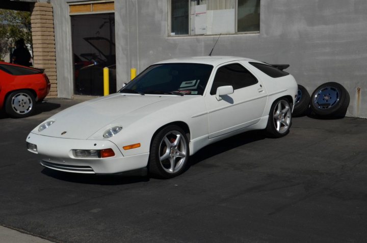 928 side skirt help - Page 1 - Front Engined Porsches - PistonHeads - The image portrays a white coupe parked in a parking space next to a building, which is presumably a garage due to the context. An additional pair of black tires is visible in the background, hinting at the variation in tire types. The car features a distinctive front grill and side mirrors, indicative of the specific model. The presence of another car and a person in the background adds to the everyday urban scene.