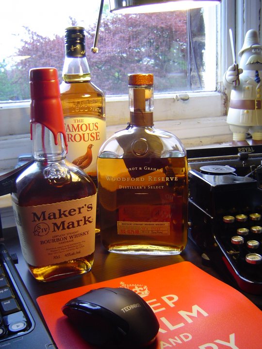 Pistonheads Official Results Election - A desktop computer keyboard is visible in the foreground with a mouse on top of it. In the foreground, there are two bottles of whisky. One bottle of whisky has a label that reads 'Maker's Mark Bourbon Whisky' and the other 'Woodford Reserve Distiller's Select'. A window with a white frame is seen behind the bottles. A red teapot and a small statue are placed near the window. A pull chain is hanging above the window. The overall setting suggests a cozy, well-organized workspace.