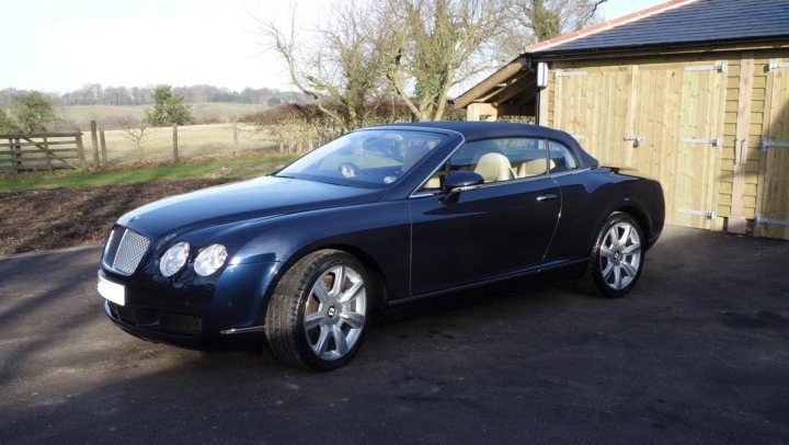 A black and white photo of a classic car - Pistonheads - The image depicts a black Bentley car parked in a gravel driveway. The car is parked facing right and appears to be a convertible model with the top down. The driveway is adjacent to a field and a line of trees. A house with a garage is visible in the background, suggesting a residential setting. The weather seems clear, as indicated by the visible blue sky.