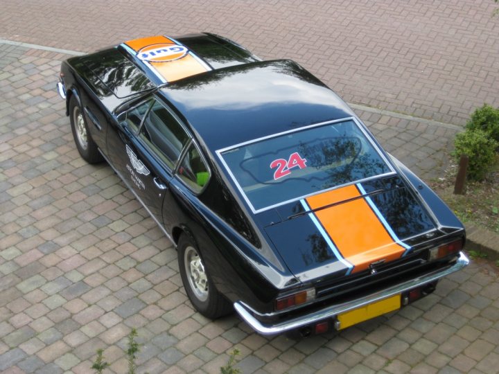 Pistonheads - The image shows a dark-colored classic car parked on a brick-paved surface. The car has some noticeable modifications, including a custom yellow stripe running across its hood, where a number "24" is prominently displayed. The car also has sport-inspired stripes on top of its roof. A circular Pirelli logo is visible on both the hood and the trunk lid. The rear portion of the car displays a license plate, and the car is positioned facing us, with the rear end to the left of the frame.