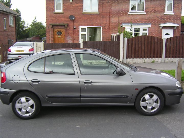 Pistonheads Bargains - The image shows a gray, four-door hatchback car parked on the right side of a street. The car is positioned parallel to the curb, which is lined with a wooden fence. A neatly maintained lawn and well-trimmed trees can be seen adjacent to the car. In the background, there are several residential houses closely spaced on both sides of the street. The sky is overcast, contributing to the generally tranquil scene.