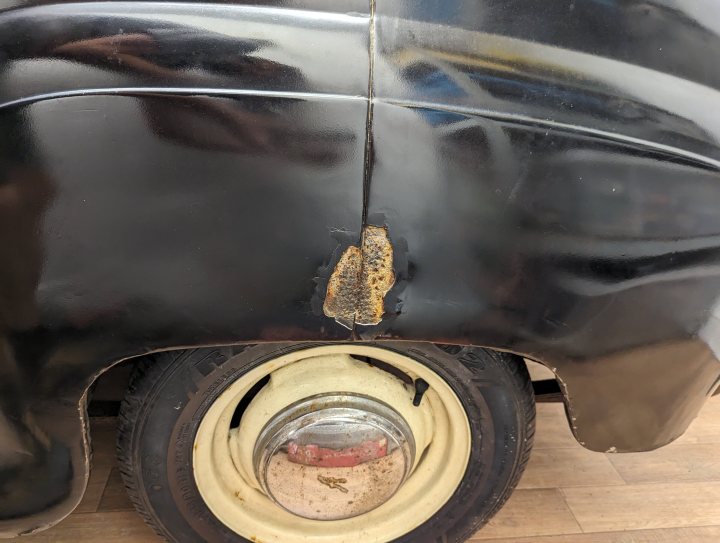 1958 Austin A35 - the family heirloom... - Page 1 - Readers' Cars - PistonHeads UK - The image shows a vintage truck with a noticeable rusted patch on the side, indicating signs of wear and possibly exposure to elements. The truck appears to be stationary, parked indoors. It has large round tires and a chrome bumper, which are typical features for vehicles from that era. The truck's overall condition suggests it has been in use or storage for quite some time. There is also a person visible in the background, but their activities or interactions with the truck are not clear due to the distance and focus on the truck itself.