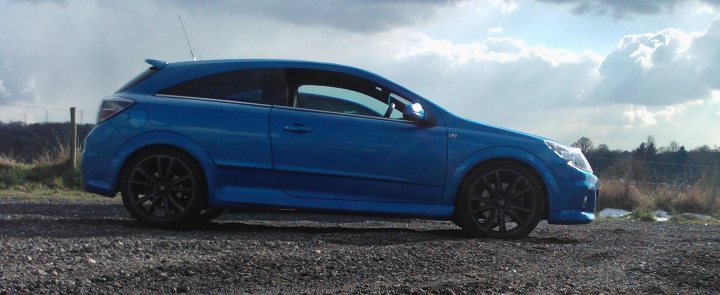 Show us your vauxhall! - Page 4 - VX - PistonHeads - The image captures a moment on a gravel road on a cloudy day. The scene is dominated by a small blue car, parked on the gravel surface. The car's position and direction suggest it might be facing out towards a large pasture area, bordered by a row of trees. The car itself is sleek and modern, with its bodywork showing a glossy shine. Despite the slightly overcast weather, the scene feels peaceful and solitary.