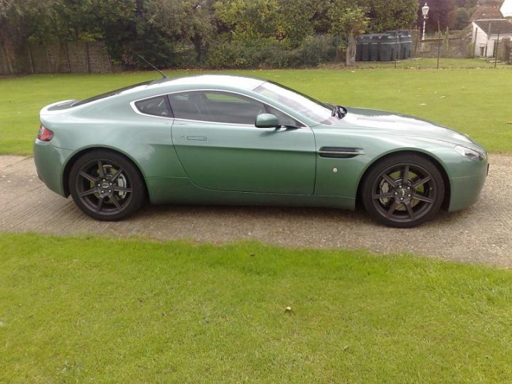 Green Pistonheads Dbs - The image showcases a sleek green sports car parked on a concrete driveway, with lush green grass visible in the background. The vehicle features a glossy metallic finish and has a distinctive side profile with a prominently contoured hood. There's a silver badge on the front, likely indicating the make or brand of the car. The interior of the car is visible through the driver's side window, revealing a typical sports car interior. There's some foliage visible near the edge of the driveway, adding a touch of nature to the scene.