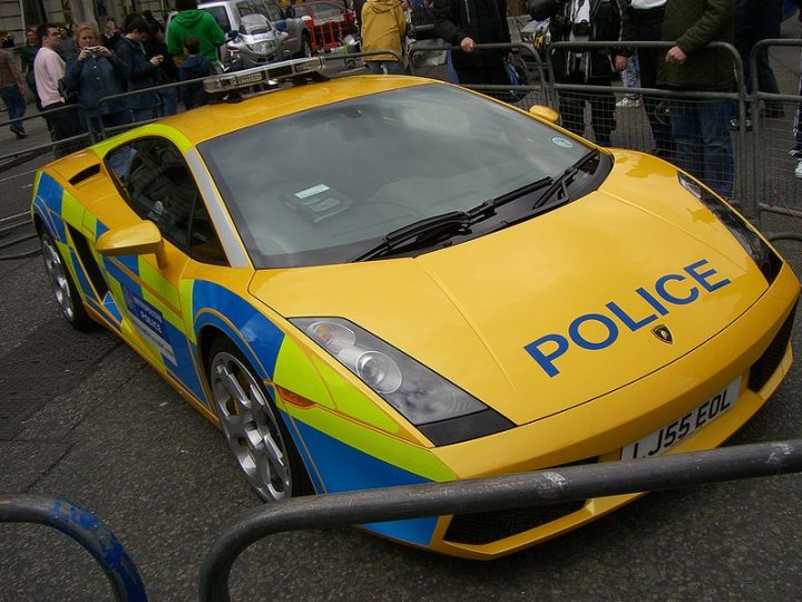 Pistonheads Dust Plod Eat - The image features a vibrant yellow sports car parked on a street. Ridged on its side is a blue and green striped print with the word "POLICE" prominently displayed. The car appears to be a Lamborghini. It's parked behind a metal barrier, separating it from onlookers, some of whom can be seen in the background. The scene seems to be a public event or spectacle, given the number of people observing the police car. The vehicle's distinctive colors and design, combined with the context of its display, suggest it is a vehicle used for police activities.