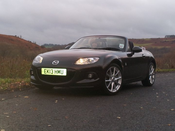 A black and white photo of a car parked in a parking lot - Pistonheads - In the image, a black Mazda Miata sports car is driving on a curvy road surrounded by hills. The car's tag says "EKI HHU." The sky is overcast, casting a soft light over the scene. There are a few trees visible on the roadside, adding to the natural beauty of the landscape.