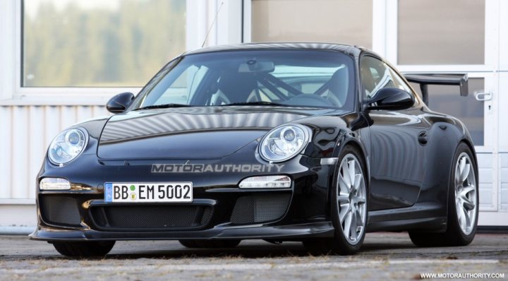 Gen Pistonheads Launch - This is a photograph of a black Porsche Cayman sports car parked on a pavement. The car is an E-Model variant with the tag B1 EM 5002, indicating it was manufactured in the year 2002. The Porsche logo is visible on the front hood, while the Renn formation, representing the number one, is displayed below the rear windshield. The car features a custom aftermarket spoiler and the body has a sleek, well-maintained appearance.