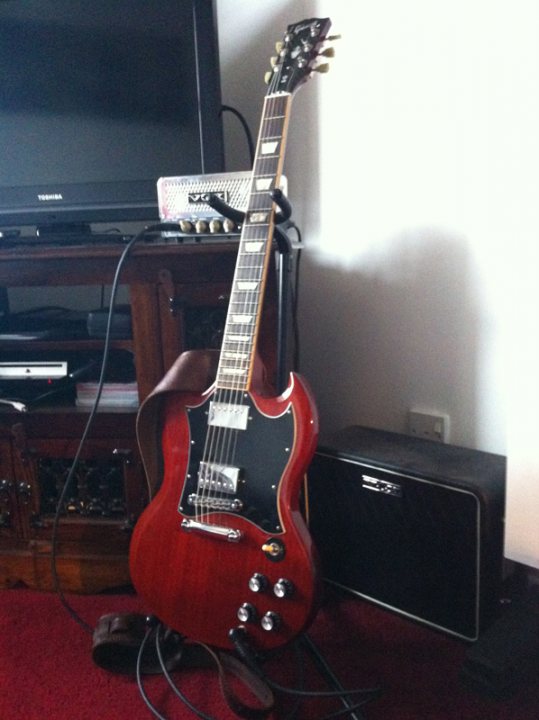 Lets look at our guitars thread. - Page 270 - Music - PistonHeads - This image showcases a wooden red electric guitar, with dual pickups, positioned upright in front of an amp, resting on a red rug. The guitar is a Fender model, known for its iconic design. To the left of the guitar, a black amp is visible, ready to provide amplification for the instrument. Behind these items, a television is mounted on the wall, providing a contrasting element to the rest of the setup.