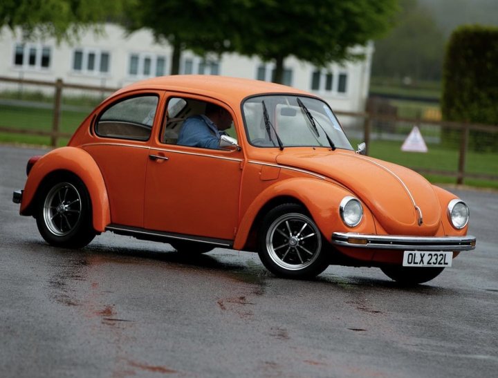 Pictures of your Classic in Action - Page 23 - Classic Cars and Yesterday's Heroes - PistonHeads UK - The image shows a classic Volkswagen Beetle car painted in a vibrant shade of orange. It's positioned on a wet, grey pavement with patches of grass visible. The vehicle appears to be moving quickly, as suggested by its dynamic posture and the splash of water behind the wheels. In the background, there are blurred images of a person and another car, indicating that this is a street scene.