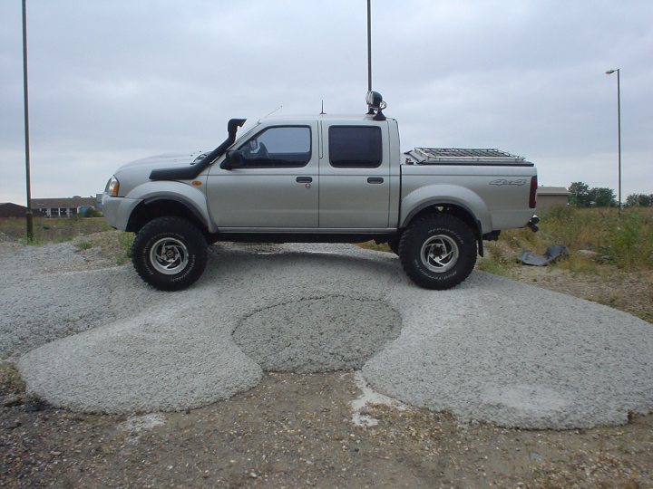 best place for Navara/Pathfinder accessories? - Page 1 - Off Road - PistonHeads - The image features a silver pickup truck with large black tires, parked atop some makeshift steps made from cement or gravel. The truck is equipped with a bed cover, and on top of this cover, you can see a small antenna. The vehicle appears to be parked in an unfinished or unpaved lot, as suggested by the rough terrain surrounding it. There is no text visible in the image.