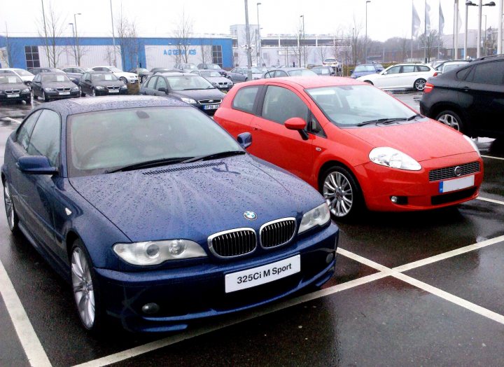 Pistonheads - The image captures an outdoor scene, where two cars are parked in a parking lot on a rainy day. The car in the foreground is a dark-colored BMW sedan, while the car in the background is a bright red Mini Cooper. Both vehicles have white license plates on the front. The scene is framed by what appears to be a store or a public parking area, with various other vehicles filling the background.