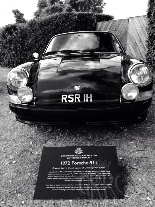 GRRC Members Annual Display - Page 1 - Goodwood Events - PistonHeads - The image shows a classic black Porsche 911, a sports car known for its performance and aerodynamic design. The car is stationary and appears to be parked outside, with a wooden fence and greenery visible in the background. Close to the front of the Porsche, there is a square plaque on the ground, which seems to provide information or a display about the car, possibly related to its history or value. The car's front head lights are prominent, reflecting a bit of the surroundings, which could suggest an overcast sky. The car's vintage design is distinct, featuring a long hood, short rear deck, and a sloping rear windshield.