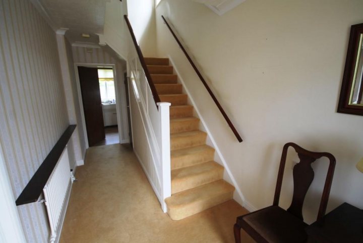 Staircase update (Easy to alter?) - Page 1 - Homes, Gardens and DIY - PistonHeads - The image shows an interior space, specifically what appears to be a hallway or stairwell. A carpeted staircase with white risers ascends to the upper level, flanked by a wooden handrail on one side. On the left, there's an open door providing a glimpse into another room, possibly a bathroom given the visible sink. To the right, a wooden chair is positioned, likely for someone to sit opposite the stairwell, but the surface it's placed on isn't visible in this shot. The door on the right side of the image, situated next to the stair railing, is closed. The walls are painted in light colors, creating a bright and clean ambiance.