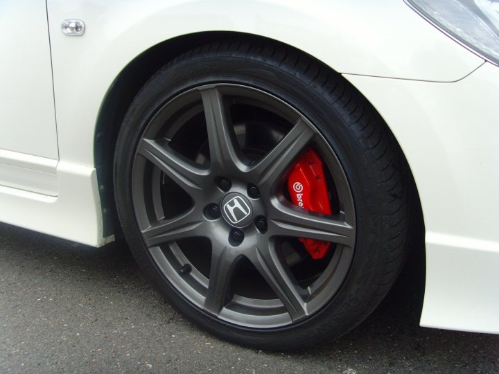 Fdr Honda Pistonheads - The image shows a close-up of a rear wheel on a vehicle, possibly a sports car, with a distinctive red Gredge Honda badge on the side of the wheel. The wheel has a five-spoke design and appears to be of a high-performance material, likely aluminum or forged alloy. The vehicle itself has a white body with a visible design element near the wheel. The settings suggest this photo was taken in an urban environment, as evidenced by the paving and the cast shadow of the car.