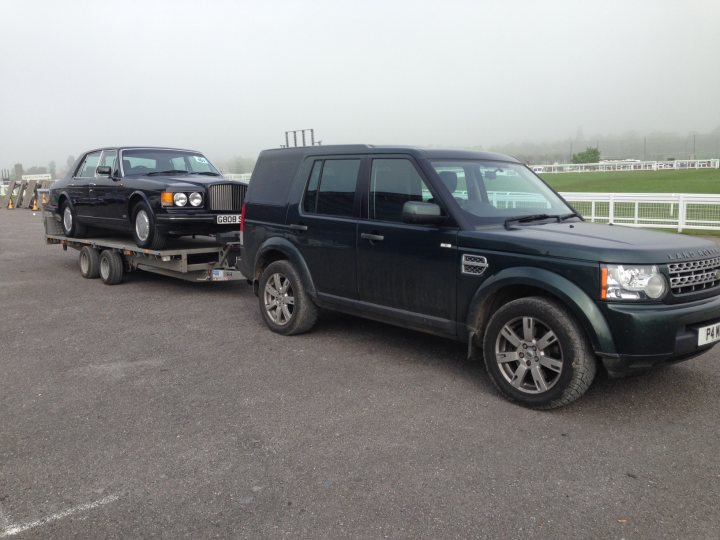 Have we been foolish? Yes but we're all grinning from ear to - Page 1 - Rolls Royce & Bentley - PistonHeads - This image features a dark-colored SUV hauling a black luxury car on a trailer. The trailer and cars are resting on an asphalt surface, and in the distance, there is a green, grassy field which suggests that this scene could be taking place in a rural or agricultural area. The SUV is depicted from the front, with its grille and headlights clearly visible. It is a clear day with no visible signs of fog or other weather conditions. The luxury car being towed is a convertible with a classic design, as indicated by its rounded front and prominent grille.