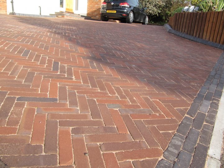 Block paving recommendations - Page 1 - East Anglia - PistonHeads - The image showcases a beautiful brick driveway. The brick driveway is positioned in front of a house and is neatly organized in a hexagonal pattern. The bricks used are predominantly brown and red, adding a rustic charm to the driveway. On the right side of the driveway, there are trees, providing a natural and tranquil backdrop to the scene. On the left side, there's a car parked, possibly indicating that someone lives in the house. The overall scene exudes a warm and welcoming ambiance.