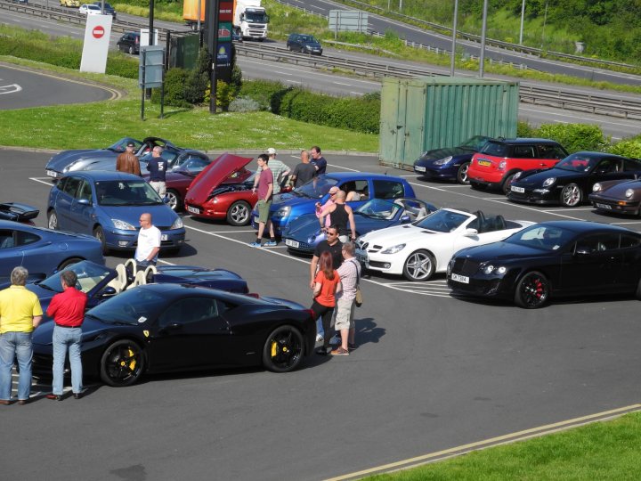 Gordano this morning en route to cwc - Page 1 - South West - PistonHeads - The image captures a bustling scene in a parking lot filled with a variety of luxury and sports cars. People are milling about, some near a black convertible, while others are gathered around a Ferrari with its hood up. There are at least half a dozen cars parked, each with its own unique design. A man stands out, standing next to a red sports car. In the background, traffic flows on a nearby road, adding to the dynamic atmosphere of the scene.