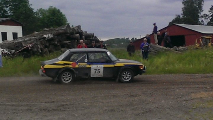 SAAB 99T rallycar pics - Page 1 - Saab - PistonHeads - The image depicts an outdoor scene of a vintage race car on a road. The car is silver and black with the number 75 on it, suggesting it may be participating in a rally or race. The vehicle is the focal point of the image, set against a backdrop of tall grass and buildings that resemble a barn and a traditional wooden shed. The sky is partially cloudy, which indicates the weather is overcast. It is difficult to discern the specific actions of the individuals due to the image's perspective, but there are several people in the vicinity, suggesting they might be spectators or crew members.