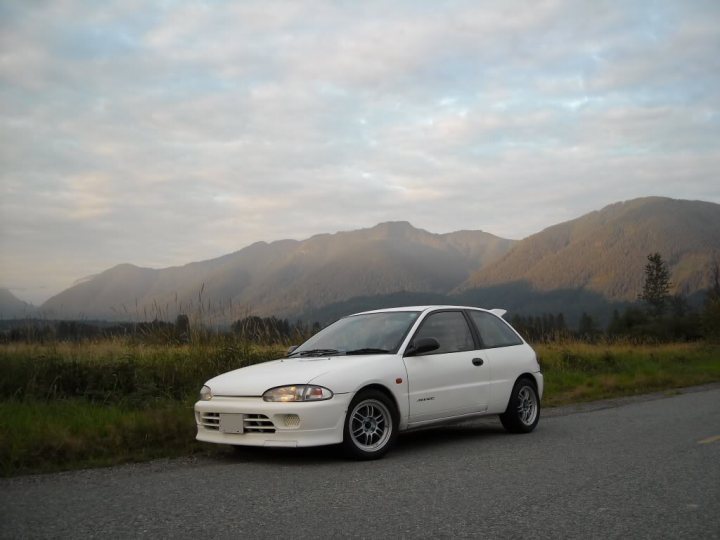 There must be a JDM car thats perfect for sprinting! - Page 1 - Jap Chat - PistonHeads - The image features a white Toyota Celica parked on a gravel road at the foot of a majestic mountain range. The car, with its polished rims and clean bodywork, appears to be in good condition. The setting sun casts a warm glow on the vehicle and the surrounding landscape, highlighting the feathery clouds in the sky and the towering mountains in the distance. The tranquil scene evokes feelings of serenity and freedom, as if symbolizing the car's democracy name.