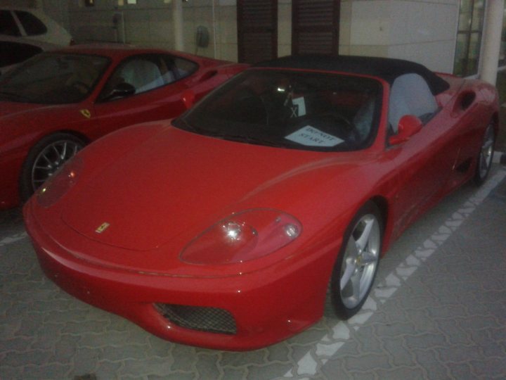 A red car with a surfboard on top of it - Pistonheads - The image shows a vibrant red sports car parked outdoors, facing the camera. The vehicle is sleek, featuring black details and a black convertible top. There's a "Top Down" sticker on the windshield, suggesting the owner is an enthusiast. The car is parked by the side of a building or possibly in a public parking lot, indicated by the white line marking its parking space.