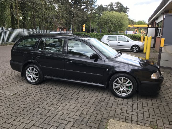 MK1 skoda Octavia VRS - £800 auction indulgence  - Page 1 - Readers' Cars - PistonHeads - The image shows a dark-colored, five-door hatchback car parked on a brick pavement. The car appears to be a compact model, possibly from the late 20th or early to mid-2000s, judging by its body shape and wheel design. The surrounding environment includes other parked vehicles, a fence, and a green tarp visible in the background. There's a pedestrian crossing in the foreground, suggesting that the car is parked at a location with foot traffic.