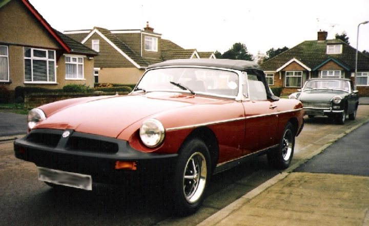 MGB or MGC? - Page 1 - MG - PistonHeads - The image depicts a classic red convertible car parked on the side of a street. The car is positioned directly in front of another vehicle, a green sedan. Both cars are parked in front of a row of houses with brown roofs. The sky is overcast, and the lighting in the photo suggests it might be early morning or late afternoon. There are no visible people or distinctive landmarks in the image.