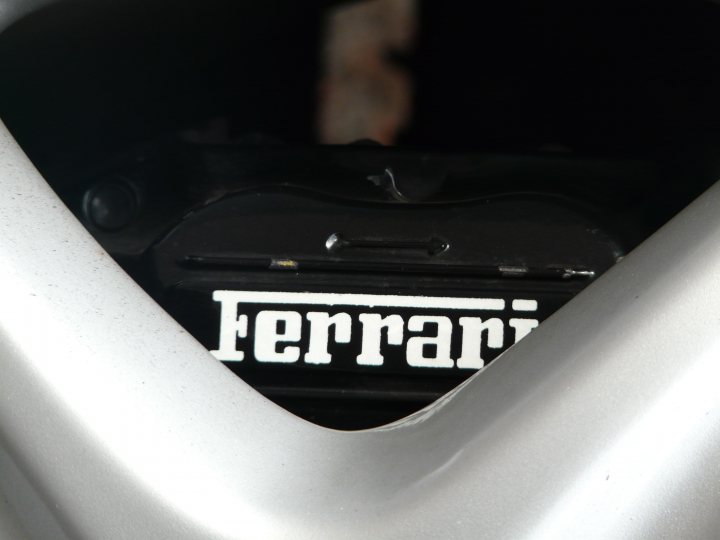 Pistonheads - The image captures a close-up of a motorcycle engine cover. The cover bears the name "Ferrari" prominently displayed across its surface. The cover appears to be black with white lettering, and it secures to the frame with a latch. The background is blurred due to the close-up perspective, but it looks like there is some kind of decorative item or motorcycle part in the background, possibly the exhaust pipe.