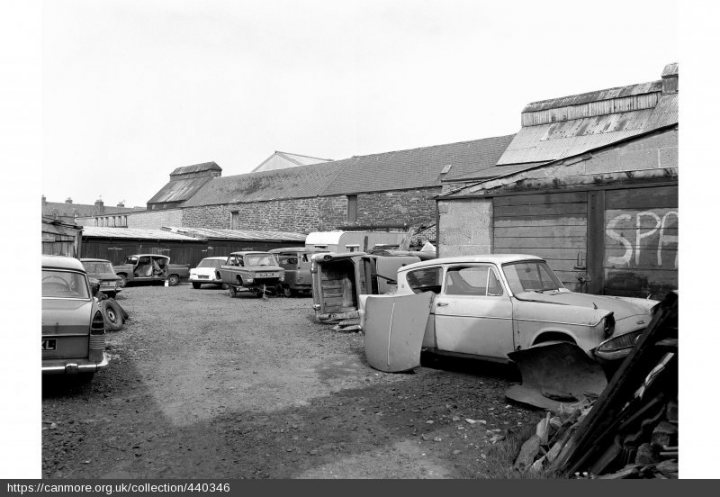 A 'period' classics pictures thread (Mk II) - Page 326 - Classic Cars and Yesterday's Heroes - PistonHeads UK - The image shows an outdoor scene featuring a collection of vintage cars parked on what appears to be a concrete surface. The vehicles range in models and colors, with some appearing more weathered than others. To the right of the cars, there is a dilapidated building with a corrugated metal roof. The setting suggests an urban or industrial area, possibly indicating a history of car manufacturing or a location once significant for its automotive industry. There are no visible people in the image.