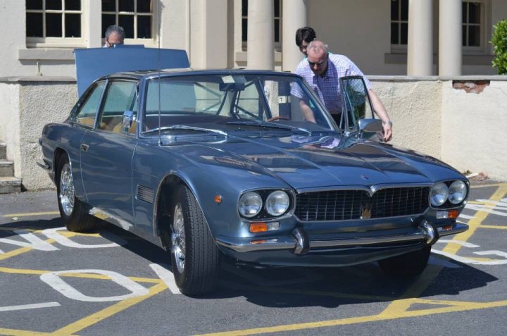 Refurbishment of my Maserati Mexico - Page 17 - Classic Cars and Yesterday's Heroes - PistonHeads - The image shows a striking blue classic car, likely a convertible model, parked on a road marked with yellow lines on both sides. The car is positioned in the foreground, occupying a large portion of the scene. In the background, two people are momentarily visible, one of whom appears to be sitting inside the car, while the other stands beside it, possibly talking to the individual inside. The car is also equipped with a pristine white hardtop, which is lifted open, revealing the interior underneath.