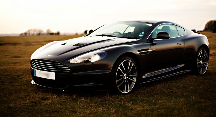 Finally had some 'pro' car shots of my DBS - Page 1 - Aston Martin - PistonHeads - The image features a modern sports car parked in an open field. The car is sleek and has a prominent wheel design. It is facing towards the left side of the photo, accentuating its aerodynamic features. The environment suggests a rural setting, with the horizon line visible in the background. A license plate on the back of the car also indicates the country it is registered in.