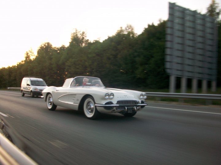 Pistonheads - The image shows a highway scene with a white vintage car driving along the right lane. The car has a distinctive design with chrome detailing and a cantilever front end. On the left lane, there's another vehicle, partially obscured, with a standard design, appearing to be a hatchback. The environment includes trees along the side of the road, indicating a rural or suburban setting, and it's captured from the perspective of the rear car. The photo is slightly blurry, giving it a painterly effect, likely taken from in-vehicle without a screen.