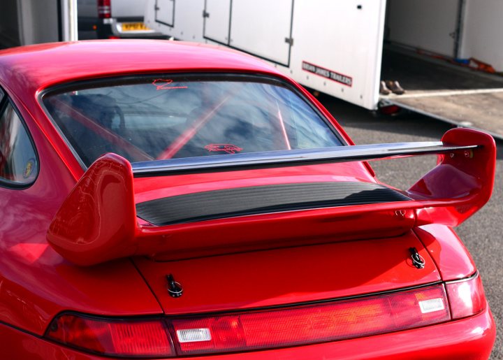 Robert Barrie Track day 24th Feb - photos - Page 1 - Goodwood Events - PistonHeads - This image features the rear end of a bright red convertible with the top down, parked outdoors. Attached to the back of the car is a Fabricated Aluminum Rear Wing (FARW), which is extended upward. Adjacent to the vehicle is a trailor with the words "FABRICATED FLAMES" and "Trailer variations" on the side, though the rest of the trailer is not visible. The scene suggests the car might be transported using the trailer. The setting appears to be a parking lot with a clear sky in the background.