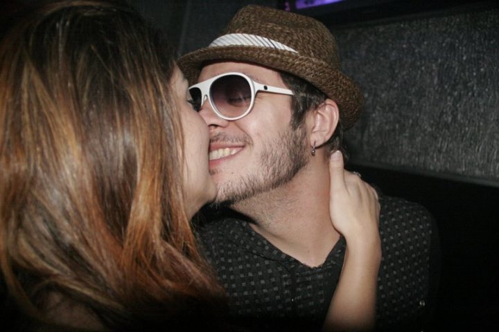 Fran Kiss - The image captures a joyful moment of two people sharing a kiss in a dimly-lit room. The man is wearing a white fedora and glasses, suggesting a playful or cool style. The woman's long hair cascades down, partially hiding her lowered eyeglasses. The atmosphere is intimate and fun, with a sense of mischief and affection in their interaction.