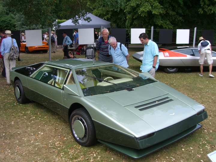 Car quiz,anyone help? - Page 1 - Classic Cars and Yesterday's Heroes - PistonHeads - The image shows a well-attended car show, likely at an outdoor exhibition space. The main focus is an older, quirky-looking green car that has attracted several onlookers. Men are engaged in conversation near the vehicle, and some carry backpacks, suggesting they might be visitors or travelers. In the background, the setting is dotted with more cars and people, contributing to the lively atmosphere of the event.