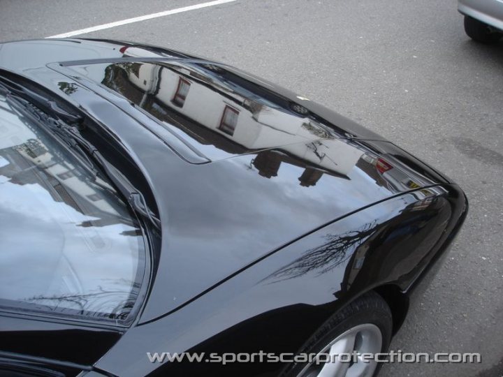 Ferrari Scuderia Pistonheads - The image is a close-up photograph of a black car's exterior section, which features the sleek back side of the car with the whiplike antenna on top. The surrounding area includes the reflection of another vehicle and buildings on the shiny paint. The photo captures the smooth texture of the car's surface and the reflection of the vehicle's front end that includes a building with a red and white striped top.