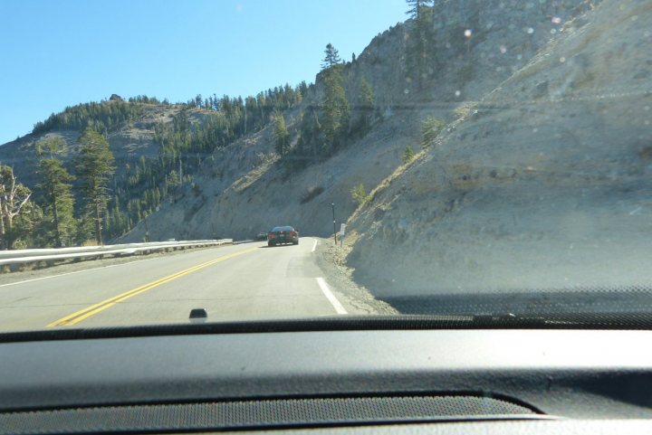 A truck driving down a highway with a mountain in the background - Pistonheads - The image is from the perspective of the driver's seat in a vehicle, showcasing a winding road cutting through a rocky landscape under a clear blue sky. The vehicle is seen from the back as it makes a curve on the right side of the road, with the edge of the dash visible. In the distance, a forested hillside can be seen, and there's a diverse mix of vegetation, with trees both in the foreground and throughout the scene. The weather appears sunny and calm, offering a panoramic view of the natural environment.