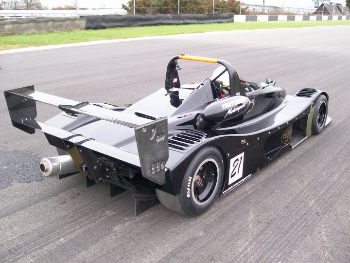 Downforcelow Downforce Pistonheads High - The image shows a sleek, black, Formula One-style race car on a track. This single-seater vehicle has prominent aerodynamic features, a large rear wing, and a prominent front splitter. The car is numbered 21 on its side, suggesting it's part of an organized racing series. The track itself appears to be a professional racing circuit with clear lane markings.