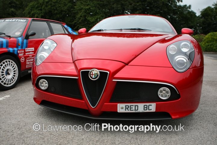 Amari Super Cars 2012 events launch photos. - Page 1 - North West - PistonHeads - The image captures a close-up view of a red Alfa Romeo car, positioned in the center. Its sleek design and shiny finish are the focal points. The license plate, which reads "RED B8", indicates a possible model or registration detail. In the background, there's another car, part of which is visible. The license plate on this car is "W51 KY". The setting appears to be quiet, possibly a parking area, with a cityscape faintly visible beyond the vehicles. The image is watermarked with "Lawrence Cliff Photography.co.uk", suggesting it was taken by a photographer associated with that website.