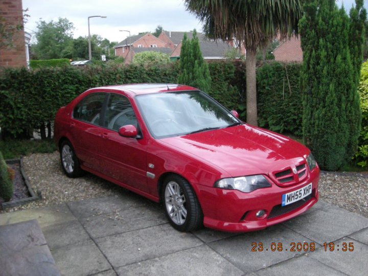 Pistonheads Diesels - The image shows a red compact car parked on a paved surface next to a well-trimmed hedge. The car is facing towards the right side of the image and is equipped with a visible license plate. In the background, there are residential buildings, suggesting that the location is a suburban area. The photo likely contains a timestamp in the lower right corner, as indicated by the date and time text overlay. The image appears to be a photograph taken during the day under clear weather conditions.