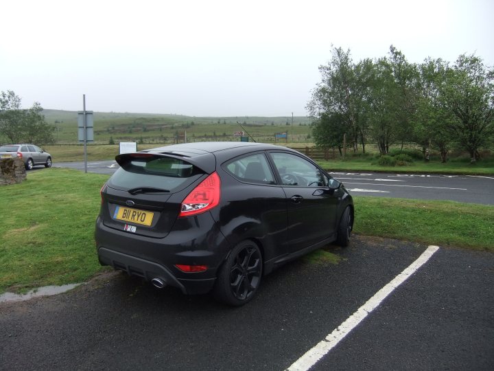 A car is parked on the side of the road - Pistonheads - The image shows a black hatchback compact car parked on the side of a concrete area marked with white lines, designated for parking. The car is facing away from the camera and appears to be parked beside a grassy area. There's a narrow road or path running parallel to the concrete area, with a few trees scattered along the side. The background is slightly blurred, making it difficult to discern specific details, but the general environment suggests a calm, open rural area with overcast skies.