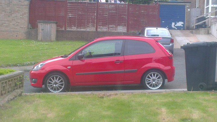 Show us your SIDE! - Page 22 - Readers' Cars - PistonHeads - The image captures a vibrant scene on a sloped street. A red car, curvaceous and stylish, occupies the foreground, parked on the side of the road adjacent to a blue trash can. The car's vivid hue contrasts with the calm presence of the waste receptacle. 

In the background, slightly atop a small hill, stands a red brick fence, adding a traditional touch to the urban setting. Beyond the fence, a green lawn slope can be seen, providing a serene and tranquil backdrop to the urban setting. 

Overall, this image portrays a harmonious blend of urban life and natural tranquility.